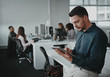 © StratfordProductions - Portrait of young businessman leaning on desk using tablet pc and his colleagues working in the background