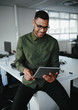 © StratfordProductions - Young businessman wearing eyeglasses sitting on the edge of office desk looking at digital tablet