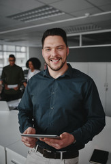 Wall Mural - Portrait of a successful smiling young businessman holding digital tablet in hand looking at camera while coworkers having conversation on background in modern office