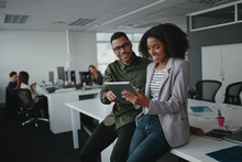 Successful Two African American Young Businesspeople Sitting On Desk Using Digital Tablet While Colleague In Background At Office 