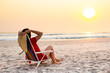 Model holding sun hat sitting on beach chair relaxing during sunset.