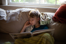 A Little Boy Resting, Reading A Book With His Blanket On A Couch