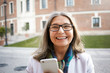 © shurkin_son - Horizontal shot of beautiful middle aged female pensioner with gray hair posing on city street, holding modern electronic device, looking at camera with happy broad smile. Age and technology