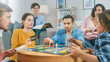 © Gorodenkoff - Diverse Group of Guys and Girls Playing in a Strategic Board Game with Cards and Dice. Man Throws Dice Impressively. Cozy Living Room in a Daytime