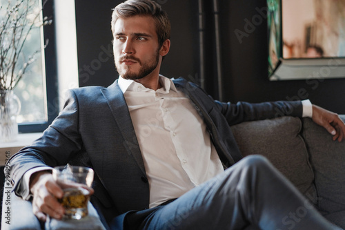 Fotografía Handsome and successful businessman in stylish suit holding glass whiskey while sitting at office