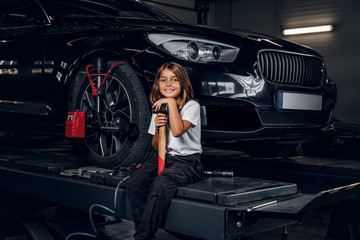 Beautiful small girl is sitting on the lifting platform with car and holding hammer.