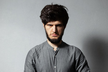 Portrait of young bearded guy with disheveled hair feels offended, on grey background.