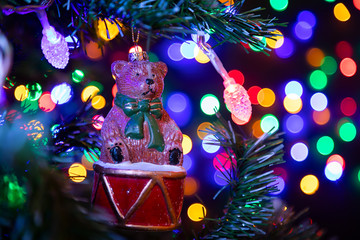 Sticker - Close-up of a shiny Christmas decoration in the form of a bear on a drum hanging on a Christmas tree in the background many garlands in different colors glow. New Year and Christmas concept.