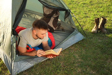 young man in sleeping bag with book lying inside camping tent