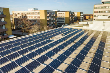 Aerial view of blue shiny solar photo voltaic panels system on commercial roof producing renewable clean energy on city landscape background.