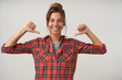 Cheerful young dark haired beautiful woman standing over white background with raised thums, pointing to herself with happy smile, wearing checkered shirt