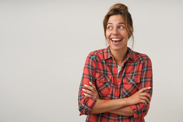 Studio portrait of dark haired happy pretty woman posing over white background in casual clothes, looking aside with broad smile and keeping hands crossed on her chest