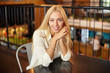 Indoor portrait of beautiful young long haired blonde woman posing over cafe interior, looking at camera with charming smile and leaning head on crossed arms