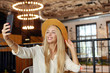 Indoor photo of charming young blonde long haired woman in brown hat posing at table in cafe, keeping mobile phone in hand and looking to camera joyfully