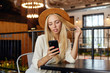 Young charming blonde woman with long hair keeping mobile phone in hand and looking at screen with calm face, wearing brown hat and white shirt