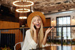 Happy young pretty blonde female having pleasant talk on phone while sitting in restaurant, smiling cheerfully and raising palm up, wearing white shirt and brown hat
