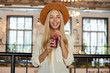 Cheerful beautiful young female with long blonde hair looking at camera happily while posing over cafe interior, holding cup of lemonade in hands