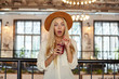 Surprised young blonde woman in wide hat posing over cafe interior, looking to camera with wide mouth opened and holding cup of lemonade in her hands