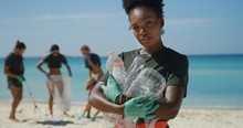 Portrait Of An Young African Female Volunteer Is Smiling In Camera Satisfied With Picking Up A Plastic Litter On A Beach With A Sea To Protect An Environment.