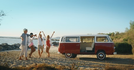 A group of young multi-ethnic friends just arrived with a minivan are happy to start their vacation and having fun on a beach with a sea on a sunset.