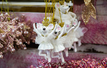 Christmas Toys In The Form Of Mice-ballerinas In White Tutus, Gold Crowns And Beads Hang In A Row In The Store, As If Dancing Together. Blurred Pink Background With Shiny Christmas