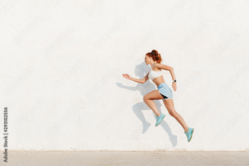 Image of redhead young woman running along white wall while doing workout in morning - obrazy, fototapety, plakaty 
