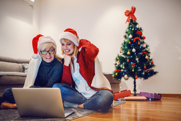 Wall Mural - Happy senior woman and her daughter sitting on floor in living room and reading messages on laptop. Both having on heads santa hats. In background is christmas tree. Family time.