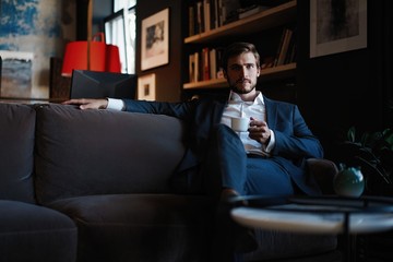 Wall Mural - Handsome young man holding coffee cup sitting on the couch in office