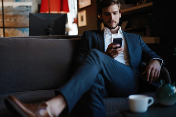 Smiling bearded man making text message on smartphone while sitting sofa at modern office.