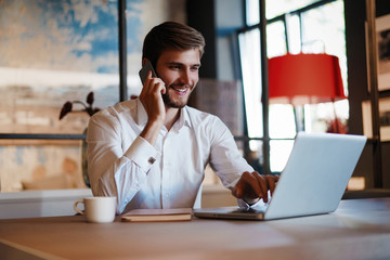 Wall Mural - Confident young man working on laptop and talking on the mobile phone while sitting at his working place in office