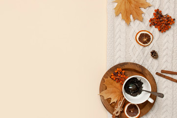 Wall Mural - Flat lay autumn composition. White cup with tea leaves, wooden plate, knitted fashion sweater or plaid, yellow maple leaves, cinnamon, dry mountain ash, orange on beige background top view copy space