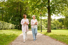 Old Age, Retirement And People Concept - Two Senior Women Or Friends Drinking Coffee Walking Along Summer Park