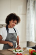 © nenadaksic - Attractive mixed race woman in apron cutting carrot and preparing dinner while standing in kitchen. On kitchen counter are vegetables.