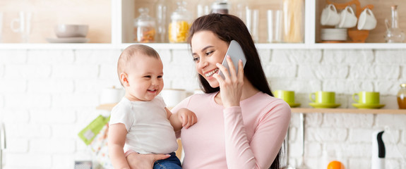 Wall Mural - Cheerful mom bonding with baby and talking on cellphone