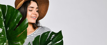 Portrait Of A Stylish Serious Girl Wearing A Straw Hat, Posing Next To An Exotic Plant. Amazing Brunette In Casual Clothes, Standing On A Light Background With Large Green Leaves