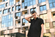 © bodnarphoto - Beautiful stylish brunette in a black shirt stands against the backdrop of a modern building with her eyes closed and poses at the camera. Attractive young male model posing on camera.