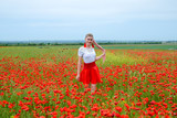 Fototapeta Kuchnia - Blonde young woman in red skirt and white shirt, red earrings is in the middle of a poppy field.