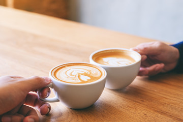 Close up image of a man and a woman clinking two coffee mugs on wooden table in cafe