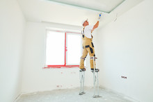 Painter In Stilts With Putty Knife. Plasterer Smoothing Ceiling Surface At Home Renewal