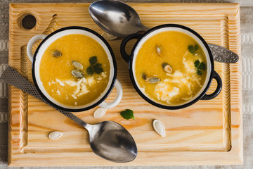 Pumpkin puree soup decorated with pumpkin seeds and cream in two bowls on a wooden tray and spoons top view