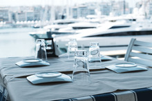 Served Table, Plates, Glasses And Frazhe In Elite Restaurant On The Seashore Against The Backdrop Of Sailing Yachts On Pier In The Marina And The Blue Sea And Sky. Table With Chairs. Tourism
