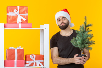 Young positive man with a beard and Santa Claus hat is standing near booth with gifts and Christmas tree on yellow background. Concept of congratulation gift and discounts for Christmas and New Year.