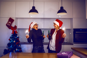 Wall Mural - Overjoyed senior woman and her daughter celebrating New Year's eve. Both having santa hats on head and beer and sparklers in hands. In background is christmas tree. Kitchen interior.