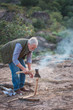 Eldery hike tourist with gray hair and beard chopping woods for campfire