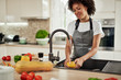 © nenadaksic - Charming mixed race woman in apron standing in kitchen ad washing yellow paprika in sink. On kitchen counter are different sorts of vegetables.