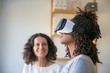 © Mangostar - Closeup of young woman in VR headset enjoying experience. Her female colleague looking at her and smiling. Augmented reality concept