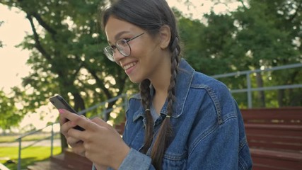 Canvas Print - Beautiful excited young brunette girl in denim jacket smiling and chatting on smartphone while sitting in the park