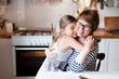 Happy mother is hugging daughter in cozy home kitchen. Woman and cute child girl are smiling. Family is using oven. Kid is enjoying kindness, embrace, care, support. Lifestyle authentic moment.