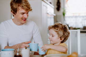 Wall Mural - Young father with a toddler son eating breakfast indoors at home.