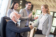 © Bojan - Beautiful middle aged blonde business woman shaking hands with older senior businessman in office. Colleagues clapping hands and celebrating successful agreement in the background.